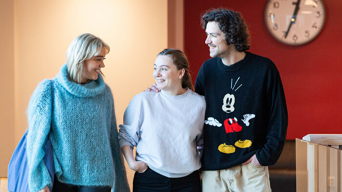 Tre smilende studenter som ser på hverandre. I bakgrunnen er det en rød vedd med en klokke på veggen. 