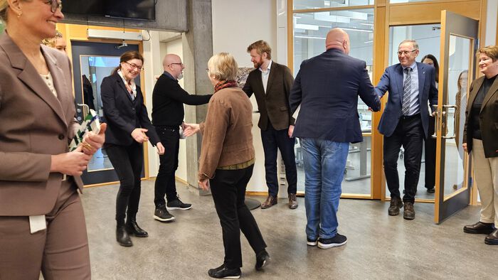 Cirka 10 mennesker møtes i en gang utenfor et møterom og hilser på hverandre. Mange smil og hender som hilser.