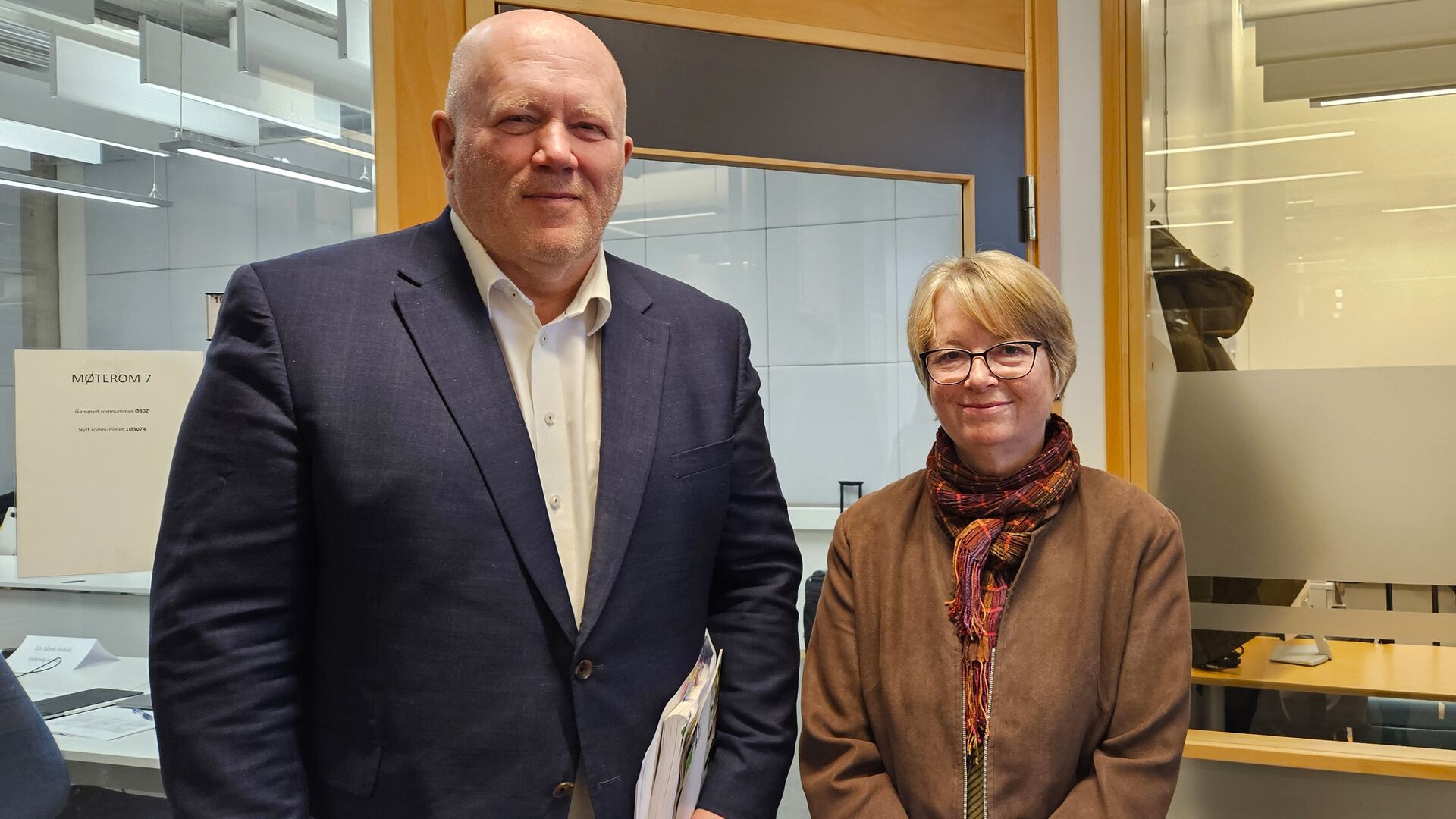 Rektor Peer Jacob Svenkerud og komitéleder Berit Johanne Kjeldstad står ved siden av hverandre og ser i kamera utenfor et møterom.