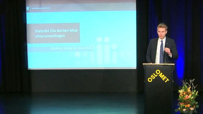 Ola Borten Moe står ved en talerstol merket Oslo Met med et powerpoint-bilde med tekst i bakgrunnen