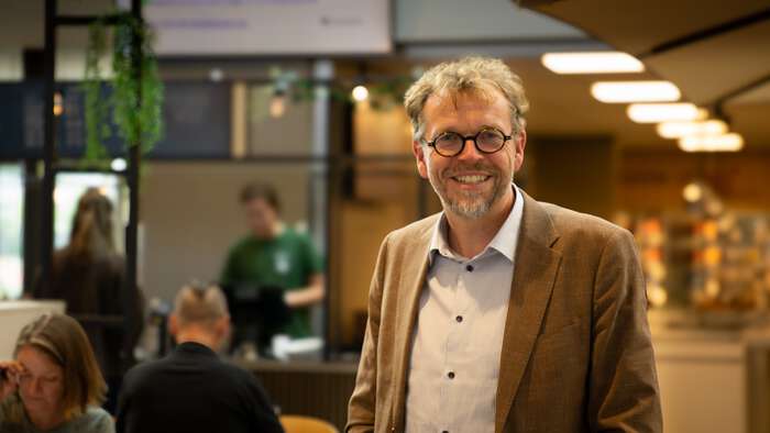 Vegard Hofsfjord står ved inngangen til kantina på studiested Elverum. Smiler til kamera. Folk i bakgrunnen.