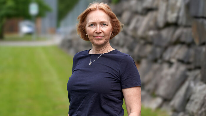 Kvinne kledt i mørke klær foran en mur og en grønn plen. 