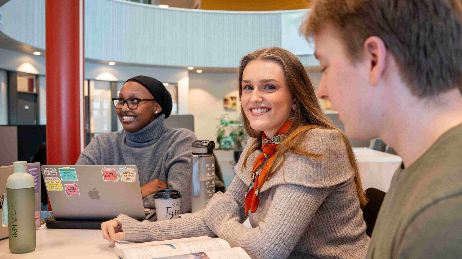 Tre studenter sitter ved et bord i biblioteket. Pc, notatbøker, vannflaske på bordet.
