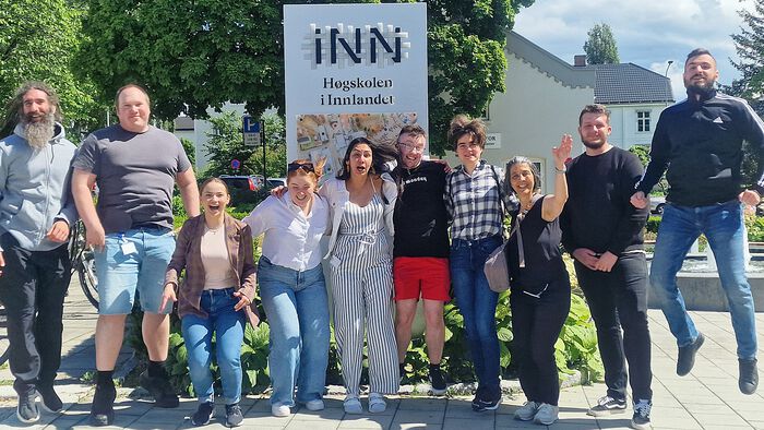 10 people are standing in front of a 'Inland Norway University of Applied Sciences' sign at Hamar campus. They are smiling and laughing.