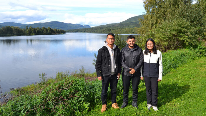Three Nepalese students are standing in front of a river with green grass and trees around them