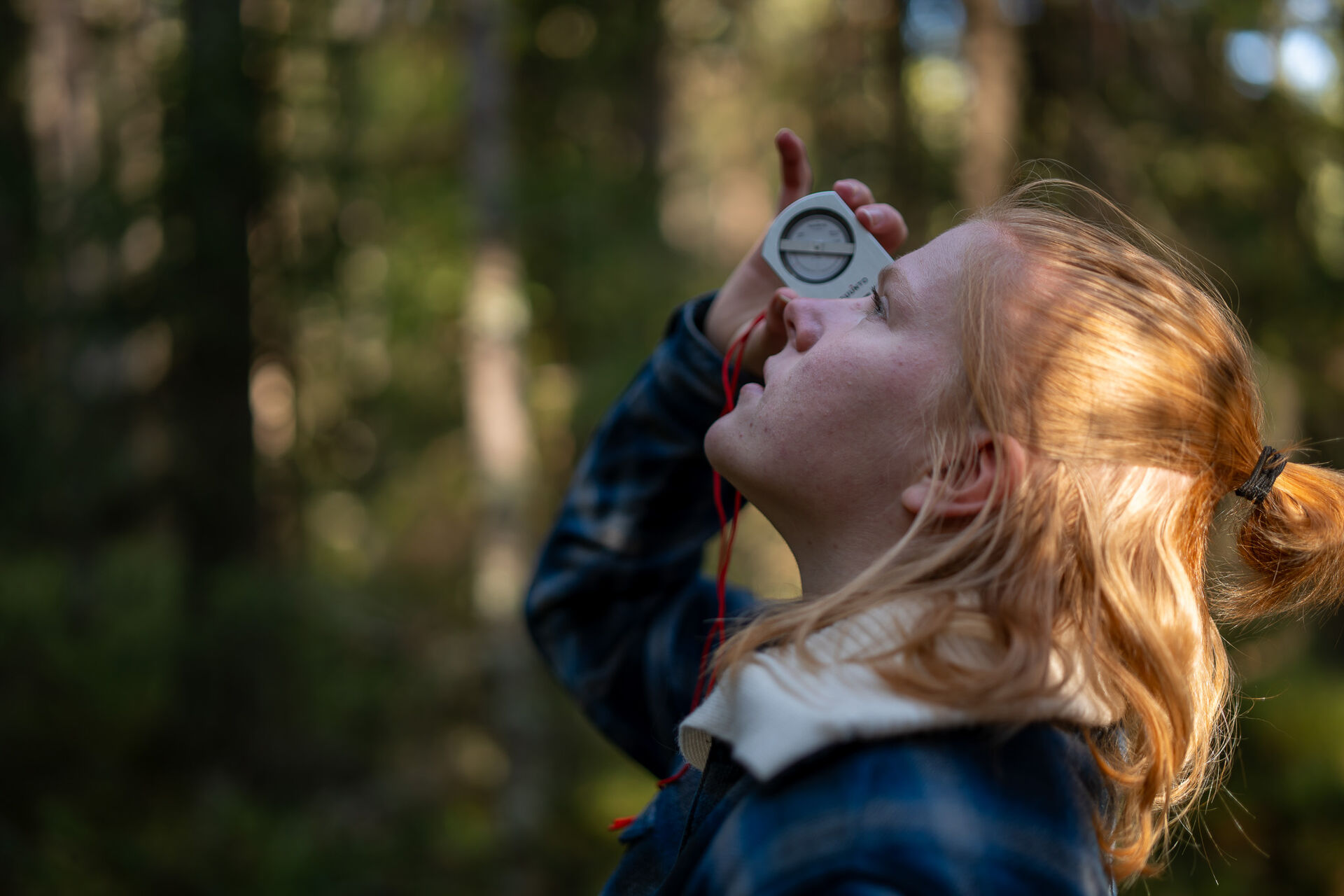 Studenten Karoline Prøis Kagnes kikker opp i luften gjennom et måleinstrument. Skog i bakgrunnen.