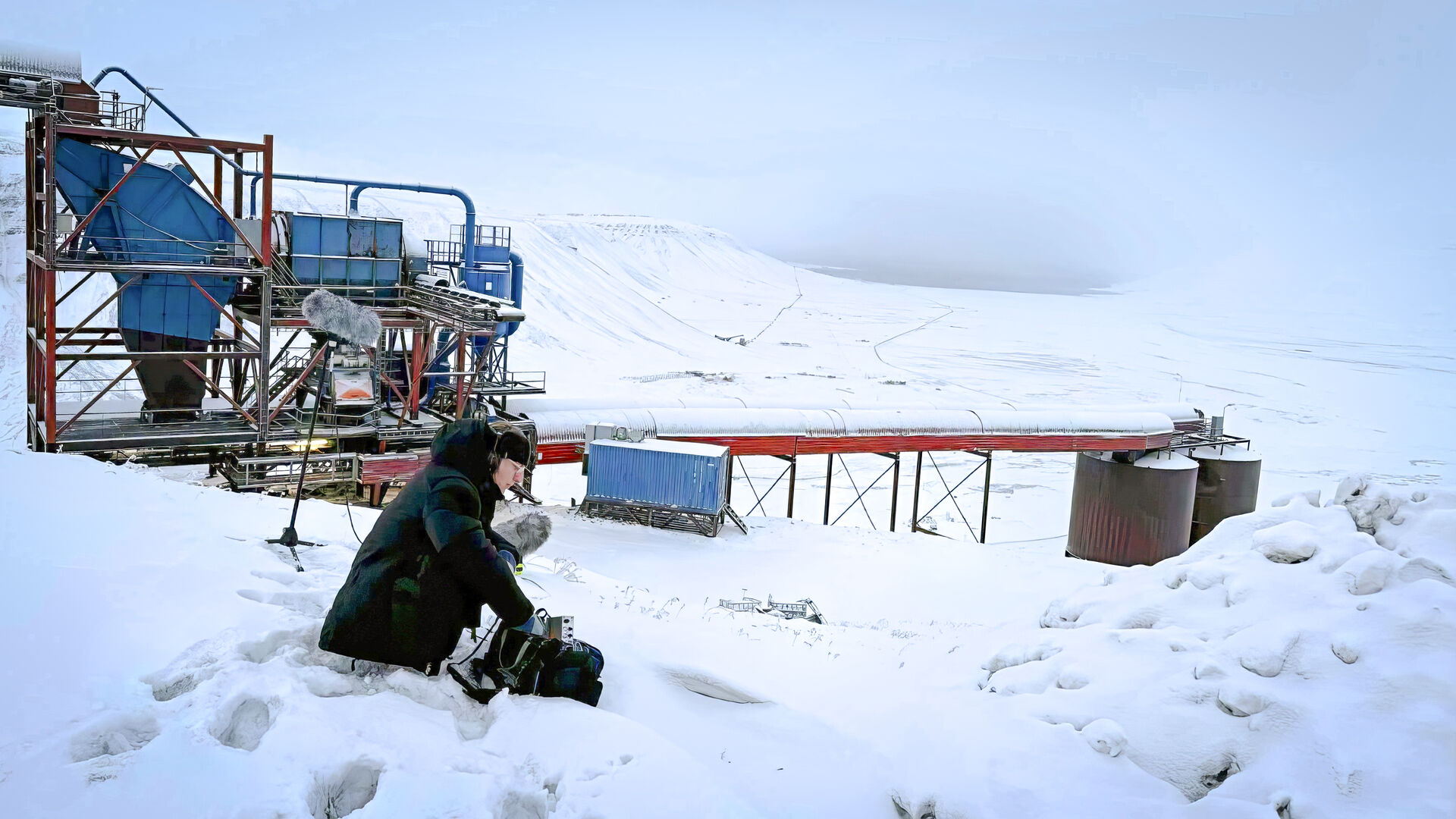 En man sitter med lydopptaksutstyr i et snøkledd landskap med en indsutriell konstruksjon i bakgrunne