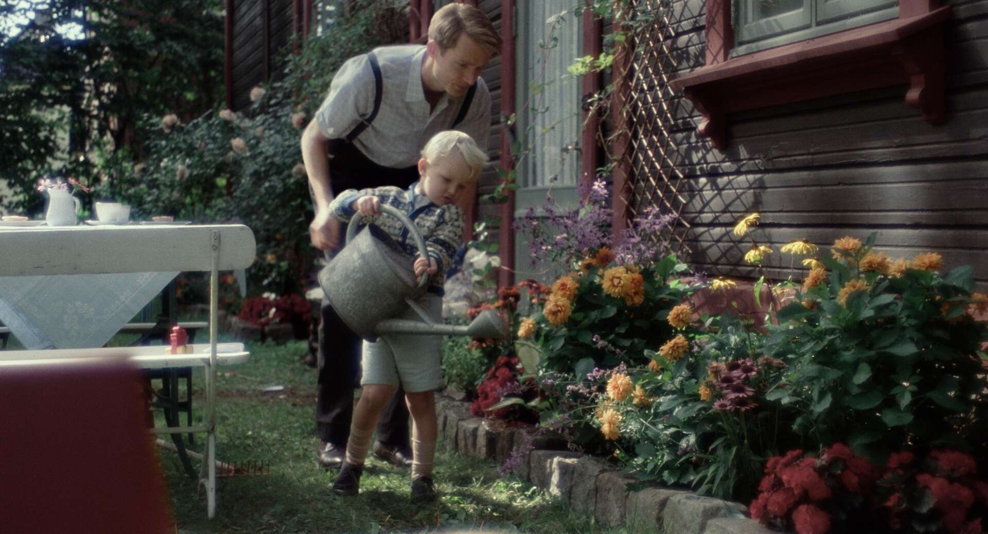 Far og sønn på 1950-tallet vanner blomster rundt husveggen.
