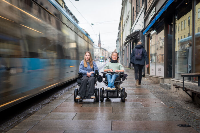 To jenter sittende i rullestol på gata i Oslo mens trikken dundrer forbi.