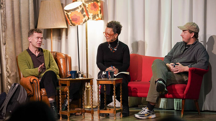Jørgen Stangebye Larsen, Eli Bø og Rasmus Eriksson i panelsamtale på scenen, som  er pyntet med en rød sofa, en brun skinnstol og tre vintage lamper.