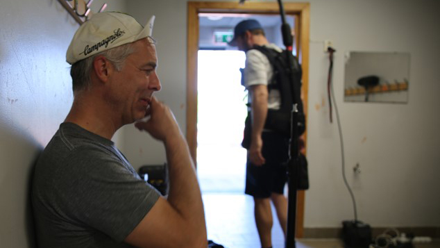 Anders Haavie sitter lent opp til en vegg mens han ser inn i en monitor. I bakgrunnen går det en mann med opptaksutstyr på seg. 