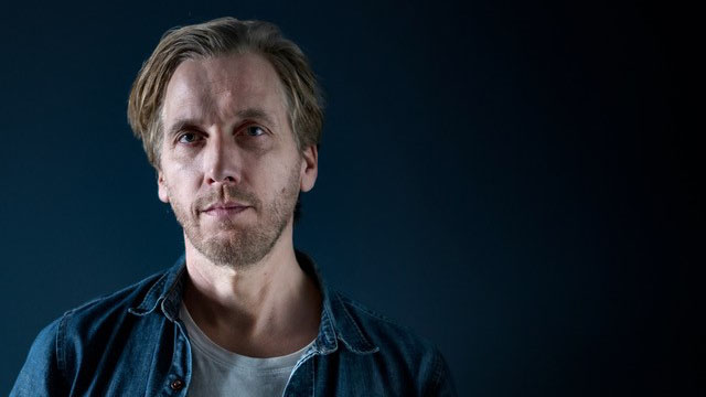 Portrett av Christian Holst Meinseth i blå skjorte mot blå, mørk bakgrunn