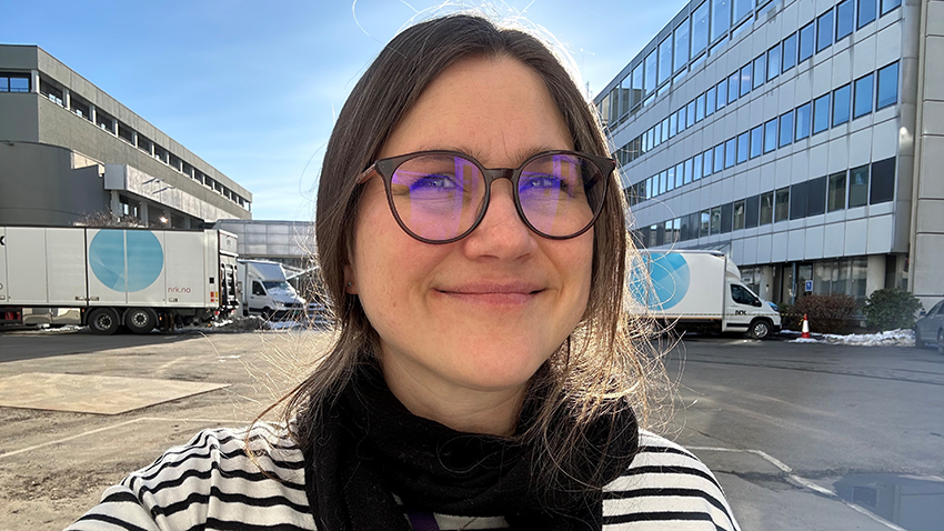 Portrett av Ingrid Løvsland i bakgården på NRK. Man skimter to lastebiler med NRK-logo i bakgrunnen. Ingrid har på seg stripete genser. 