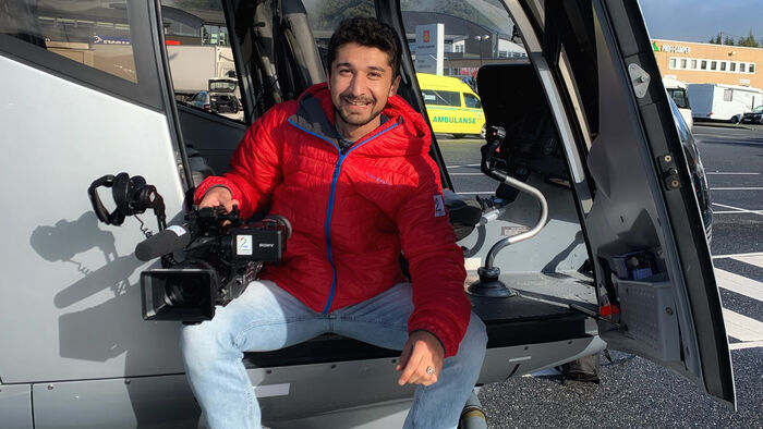 Sorosh Sadat sitter i døra på et helikopter med et kamera på det ene kneet. Han smiler og har på seg en rød jakke. 