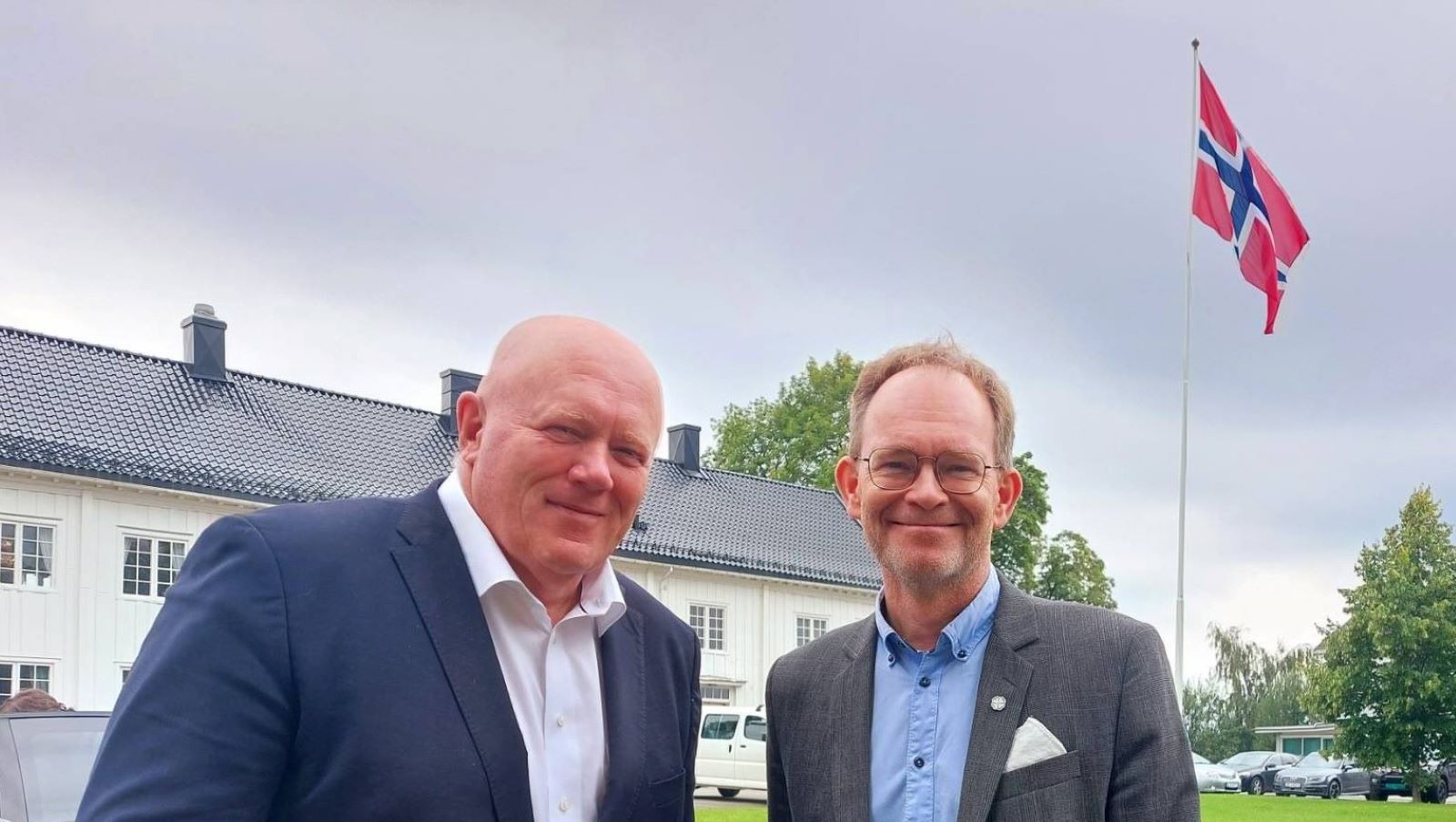 Rektor og Forsknings- og høyere utdanningsministeren smiler mot kamera med en hvit bygning og et norsk flagg på flaggstang i bakgrunnen