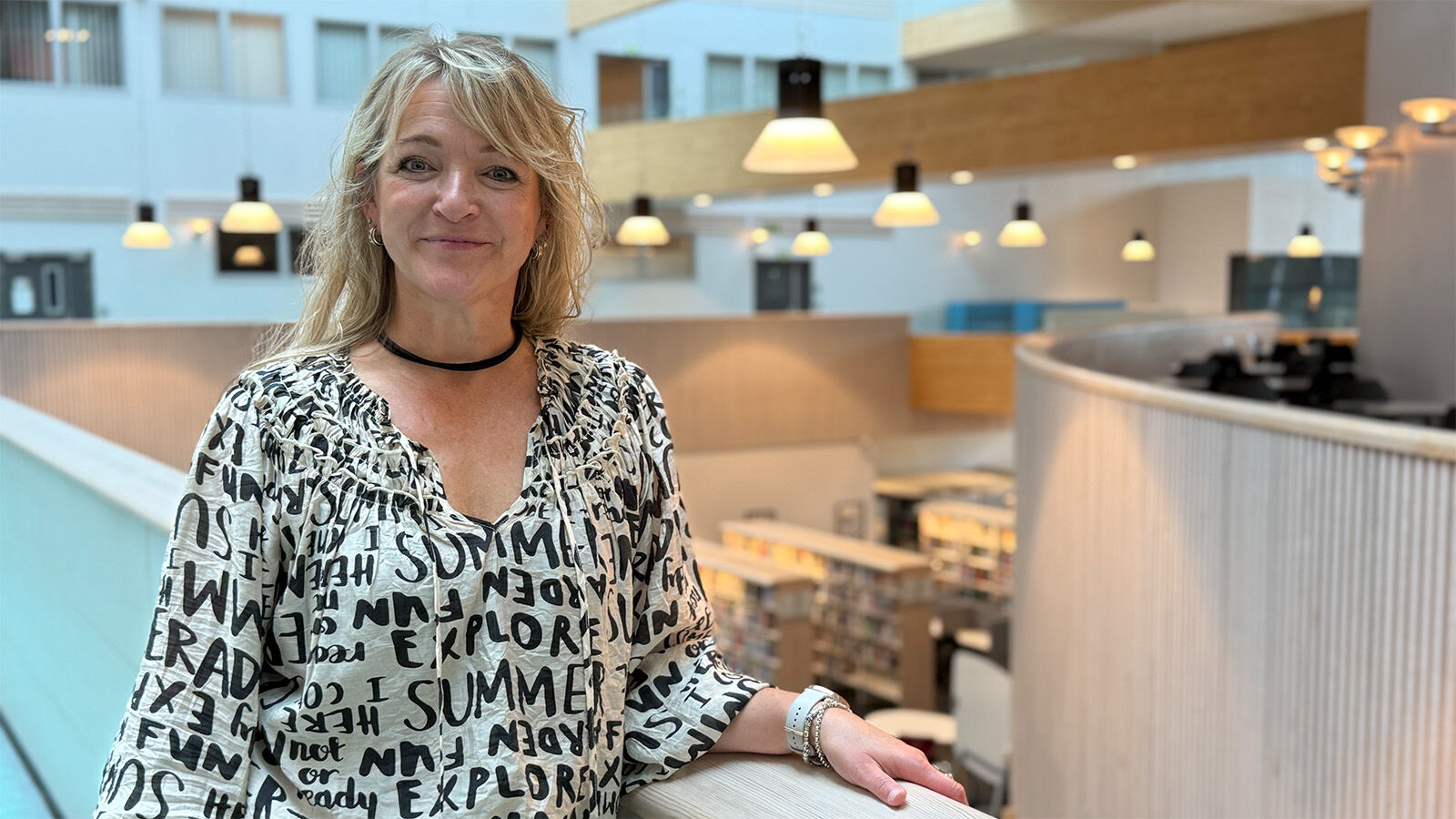 Kvinnelig ansatt står smilende ved gelender inne på studiested Elverum, med biblioteket nede i bakgrunnen