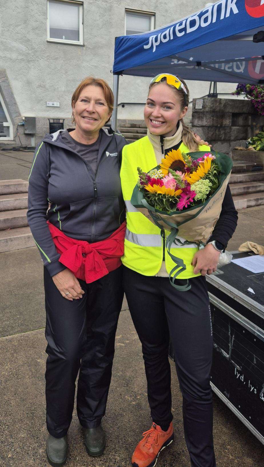 To kvinner står smilende utendørs foran et blått telt. Den yngre kvinnen, som er student Benedicte, har på seg en gul refleksvest og holder en stor blomsterbukett med blant annet solsikker, mens den andre kvinnen har mørke treningsklær og en rød genser knyttet rundt livet.