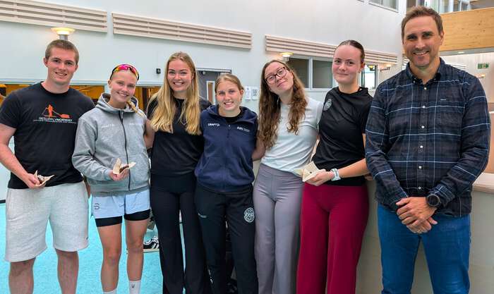 Sju studenter og instituttleder oppstilt på rekke. Noen spiser kake, alle smiler.