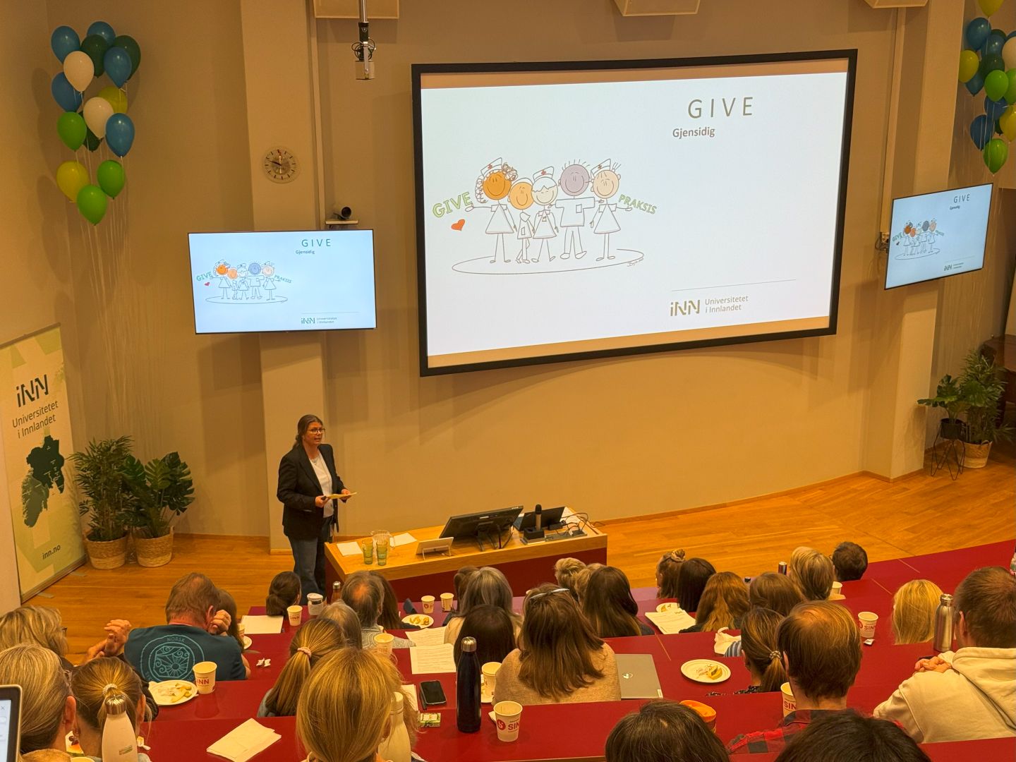 Kvinne holder foredrag med en stor skjerm med grafikk av en gjeng med fornøyde sykepleiere og sykepleierstudenter og teksten ''Give praksis" 