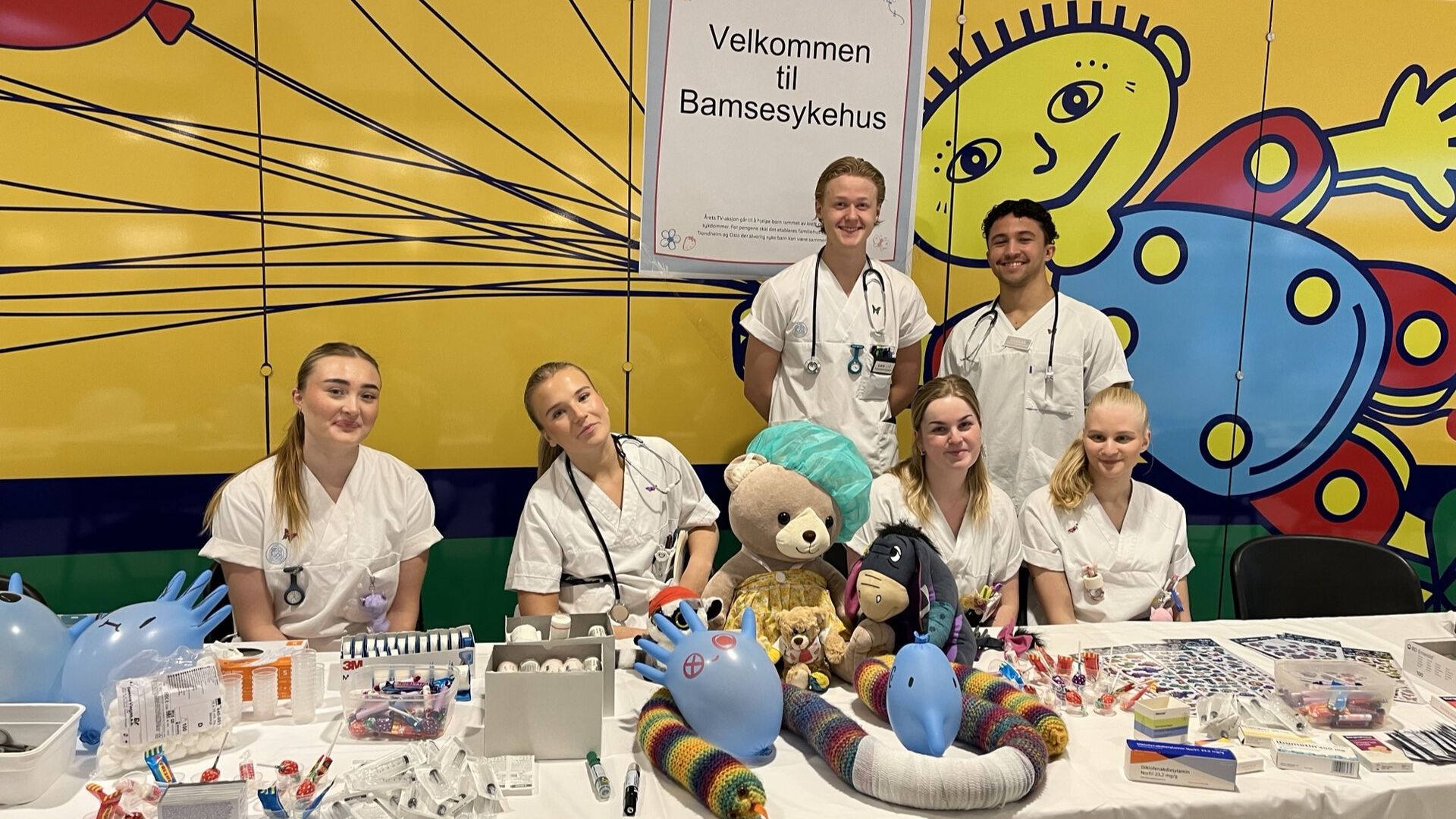 Seks sykepleierstudenter i uniform foran et langbord fylt av sykehusrekvisitter, godterier, samt et herlig knippe skadeskutte kosebamser. Foto: Barnekreftforeningen.