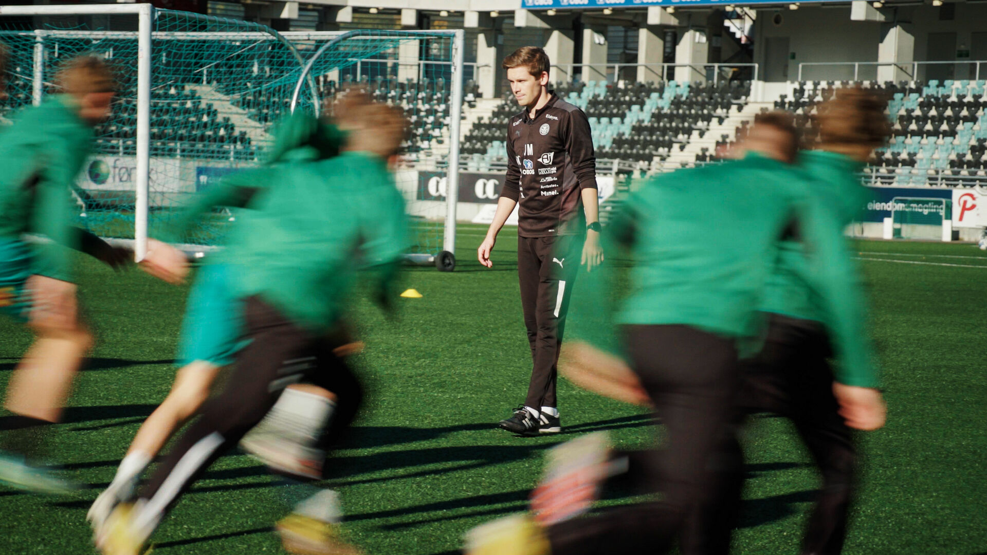 Mann står på en fotballbane mens spillere løper forbi han