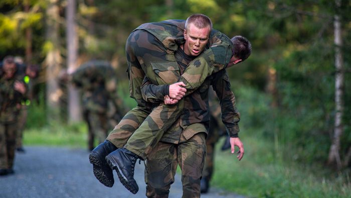 En soldat bærer på en annen soldat langs en vei i skogen, flere soldater i bakgrunnen.
