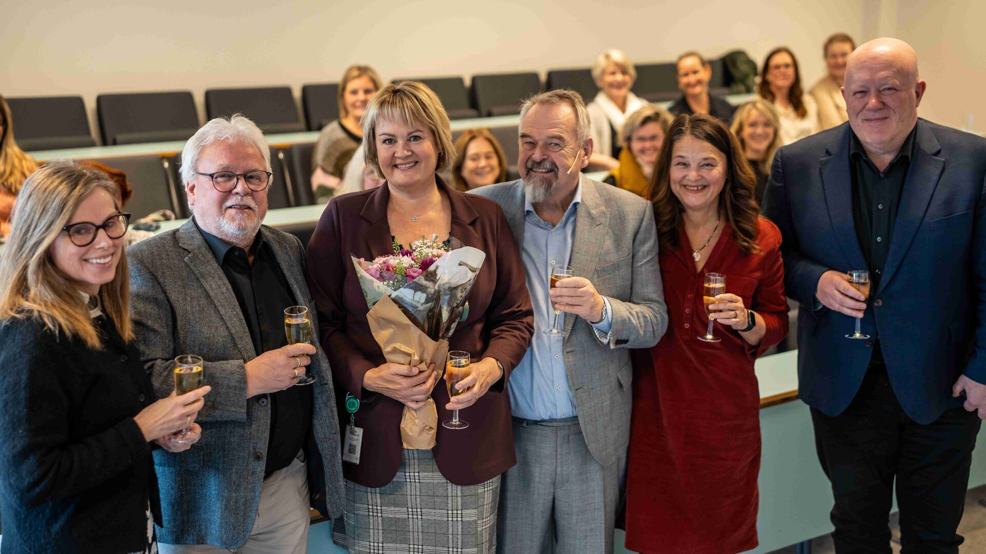 Seks personer står på rekke, hovedpersonen i midten holder blomster, alle holder et glass. Publikum sees på rader bak.