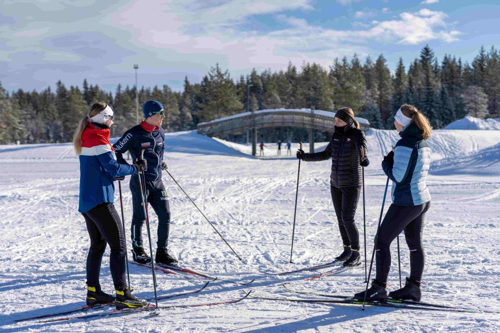 Fire skiløpere som står å snakker sammen. De har på treningstøy og det er snø på bakken.