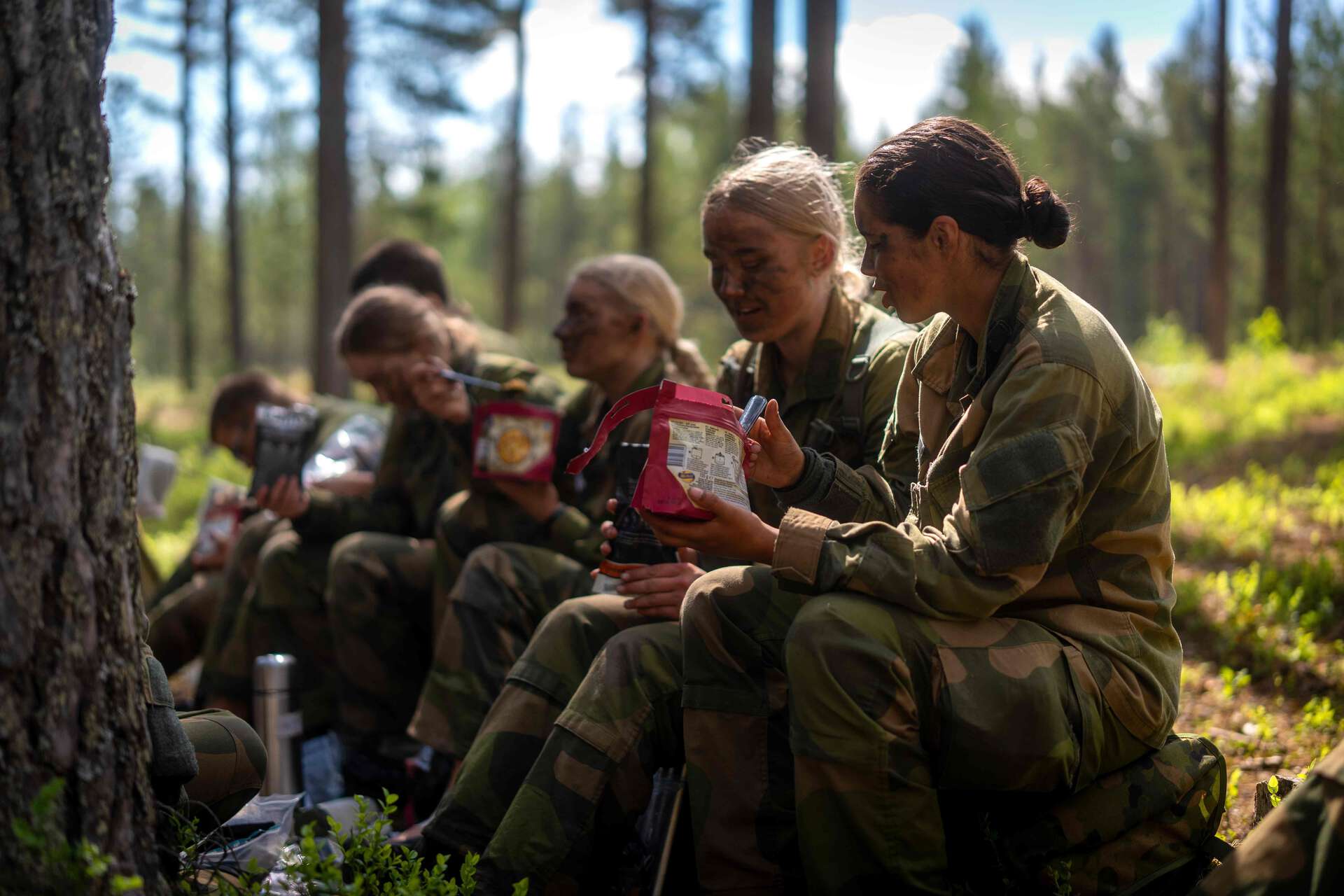 En gruppe soldater i kamuflasjeuniform sitter på bakken i skogen og tar matpause mens de holder og åpner feltrasjoner.