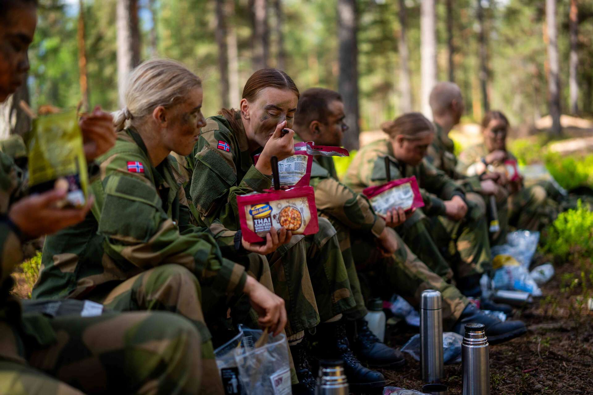 En rekke soldater i kamuflasjeuniform sitter på bakken i skogen og spiser feltrasjoner, mens poser med turmat og termosflasker ligger foran dem.