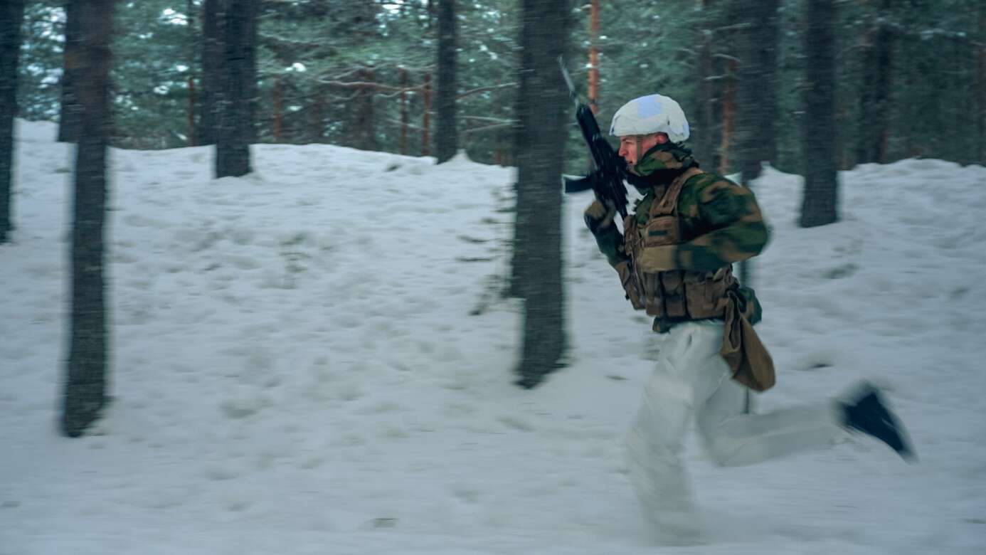 En soldat i vinterkamuflasje og hjelm løper gjennom en snødekt skog med gevær i hendene.
