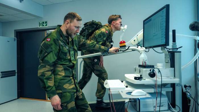 To norske soldater i uniform gjennomfører fysisk belastningstest på tredemølle i et laboratorium, mens måleutstyr og dataskjermer registrerer pust og yteevne.