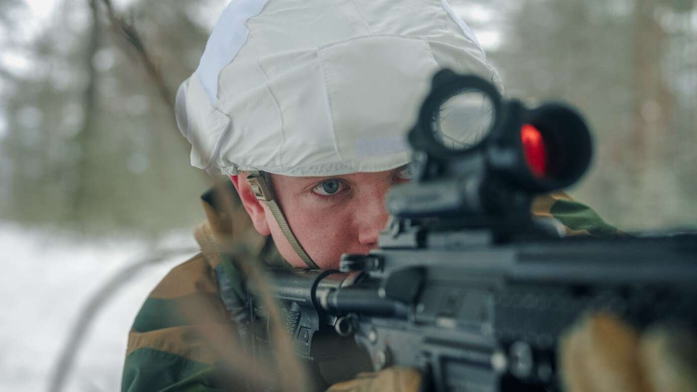 Soldat i vinterkamuflasje ligger i snødekt terreng og sikter gjennom rødpunkt­sikte på gevær under øvelse i skogen.