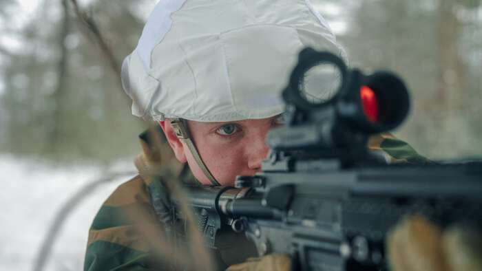 Soldat i vinterkamuflasje ligger i snødekt terreng og sikter gjennom rødpunkt­sikte på gevær under øvelse i skogen.