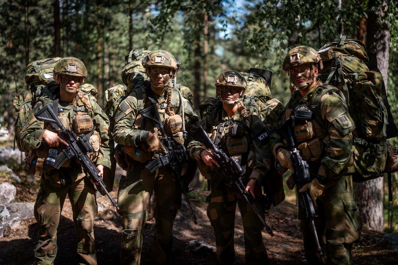 Fire soldater i feltuniform står i skogsterreng med full oppakning og våpen, klare til oppdrag under øvelse.