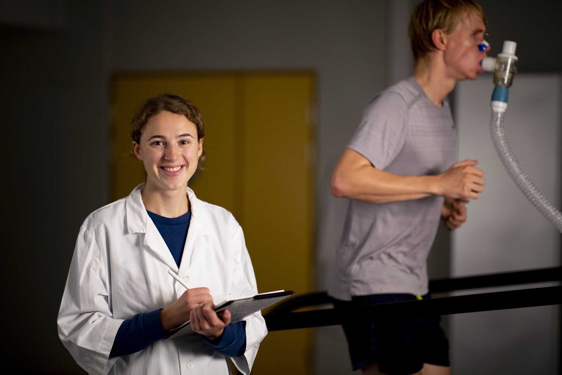 Mannlig idrettsstudent løper på tredemølle mens kvinne i hvit labuniform smiler og skriver resultatene