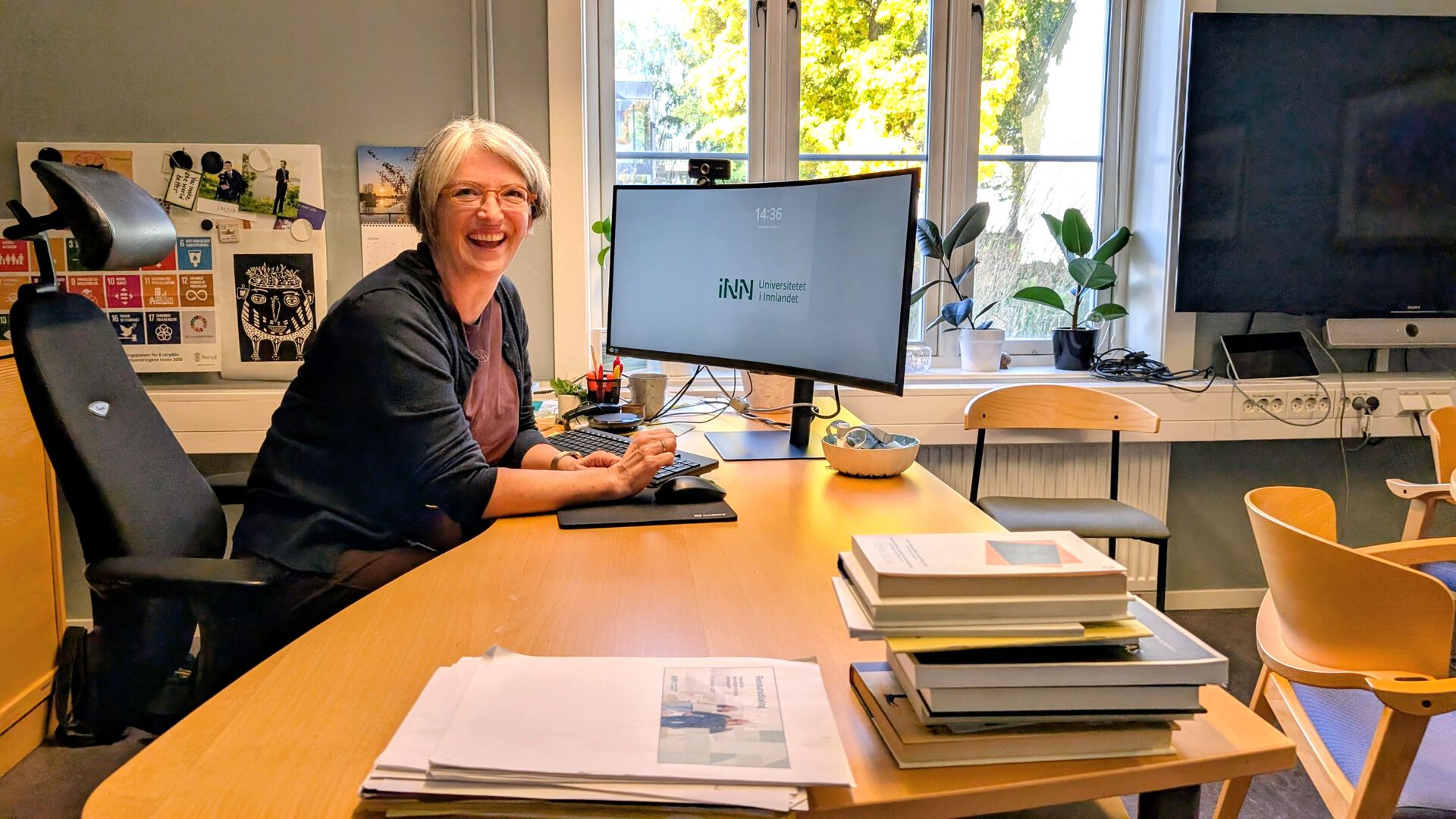 En smilende Gunhild Tomter Alstad på kontorstolen sin med utsikt til møteplassen på kontoret, til dataskjermen og ut vinduet på studiested Hamar.