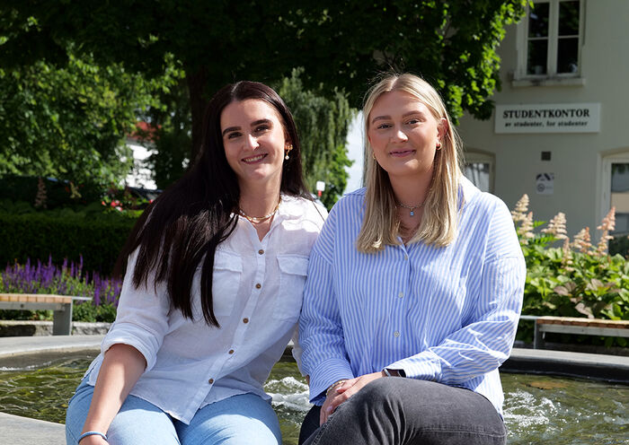 Bilde av leder for Fadderuka Andrea Lien-Ingvaldsen og nestleder Ingrid Bardosen Aanerud sittende foran en vannfontene på campus Hamar.