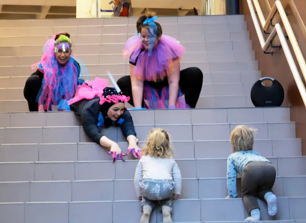 Bilde av tre studenter i trappa på høgskolen, utkledd som vetter med slør og svarte drakter, og to små barn som er på vei opp trappa for å se komme tettere på.