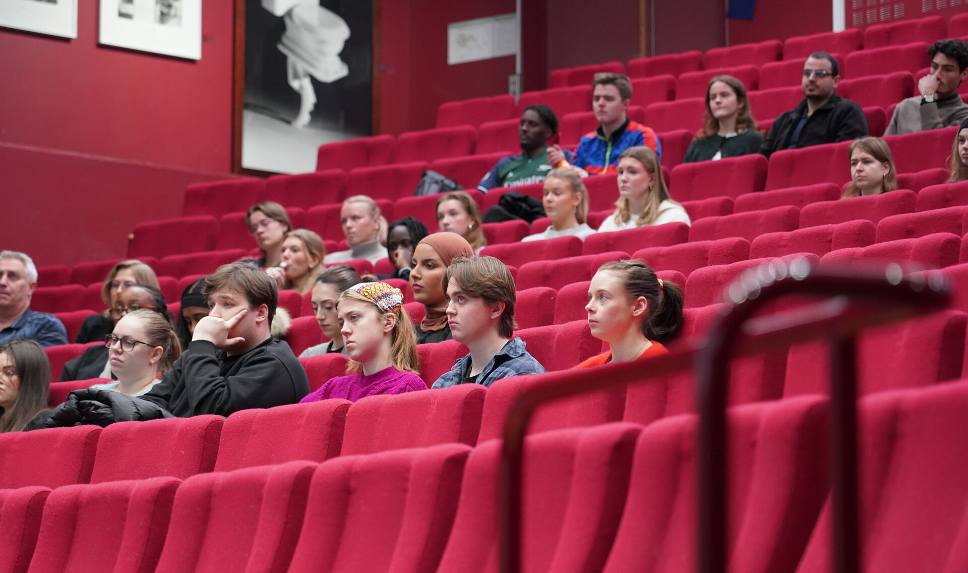 Studenter sitter i et auditorium med røde stoler og hører på et foredrag