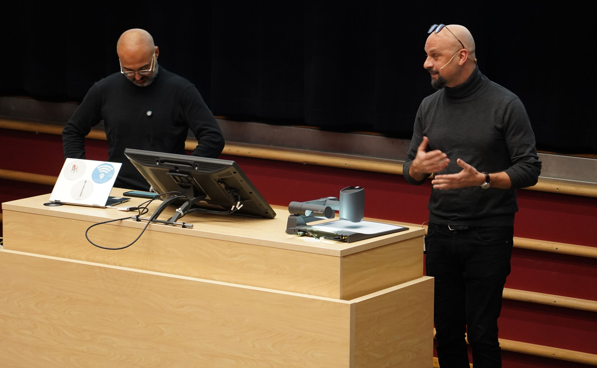 To menn i svart genser og svarte bukser står foran en svart vegg i et auditorium og snakker
