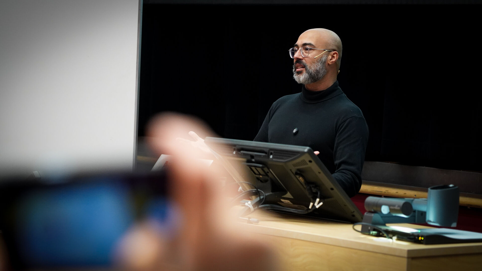 Mann med svart genser og briller står foran en svart bakvegg i et auditorium