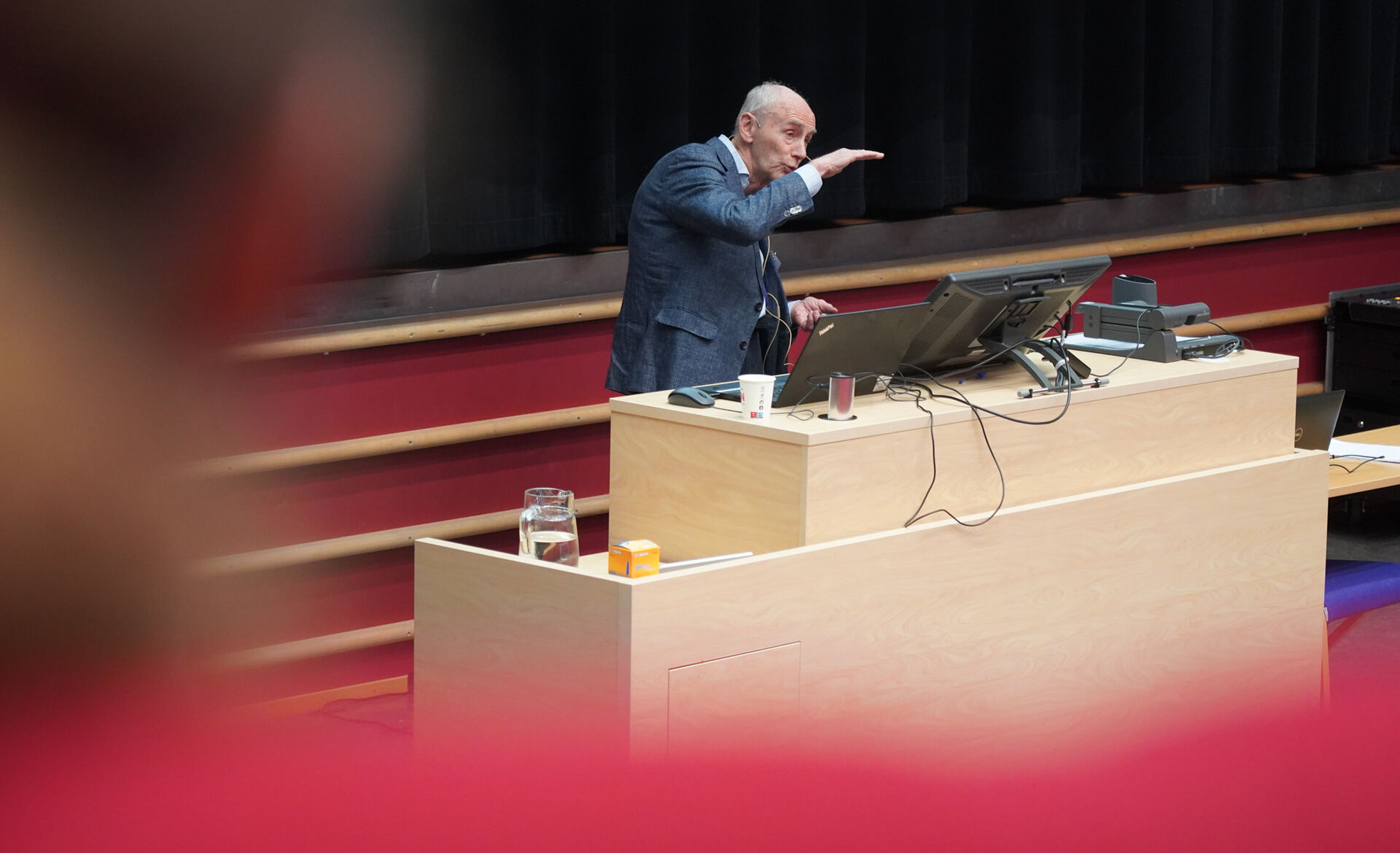 Eldre mann med grå jakke holder foredrag i et stort auditorium