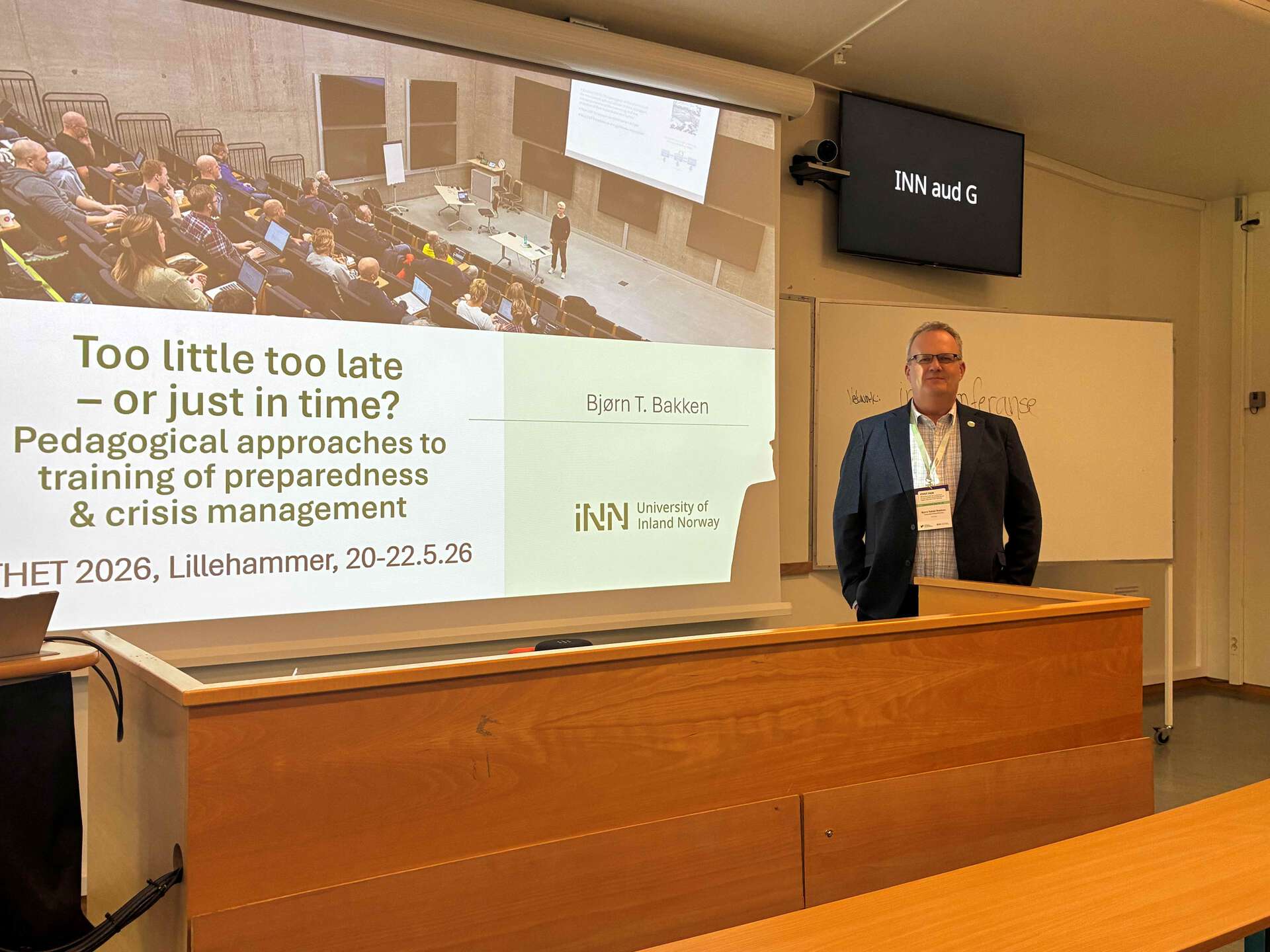 A man wearing glasses and a black blazer looking at the camera while standing on stage facing the auditorium with a slide behind him with the caption Too little too late – or just in time? Pedagogical approaches to training of preparedness & crisis management