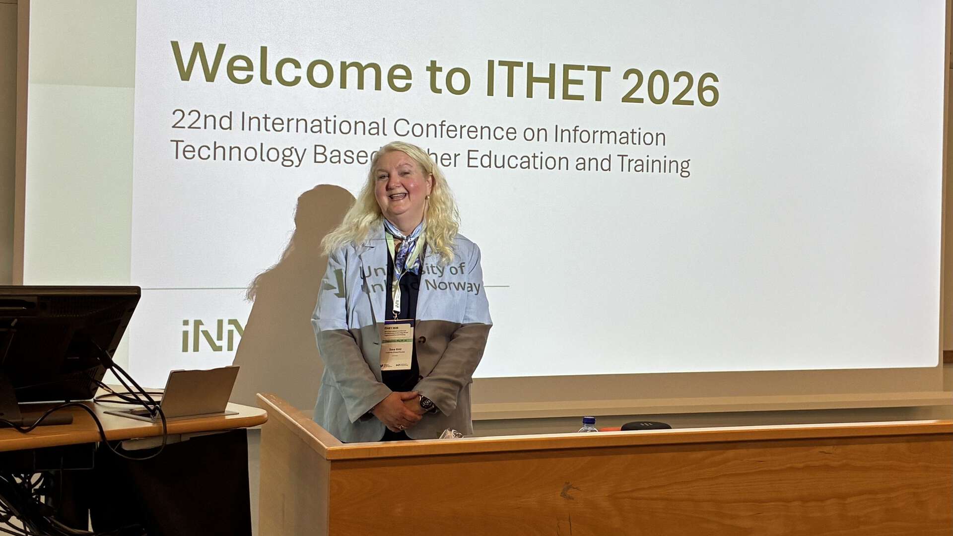 A smiling blonde woman standing at the front of an auditorium in front of a screen with the writing Welcome to ITHET 2026