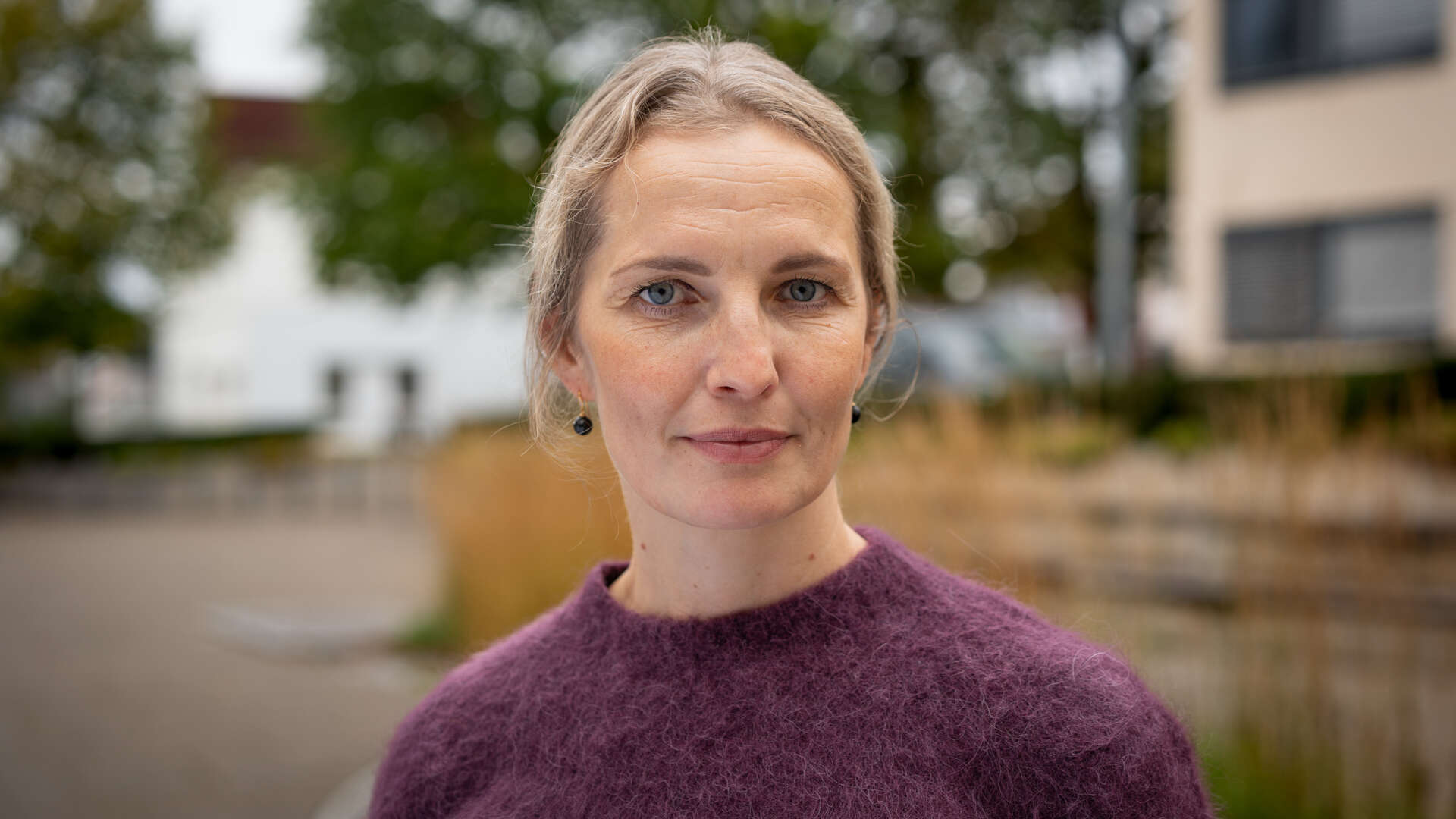 Dame med lilla genser og mørkt blondt hår med bygninger i bakgrunnen