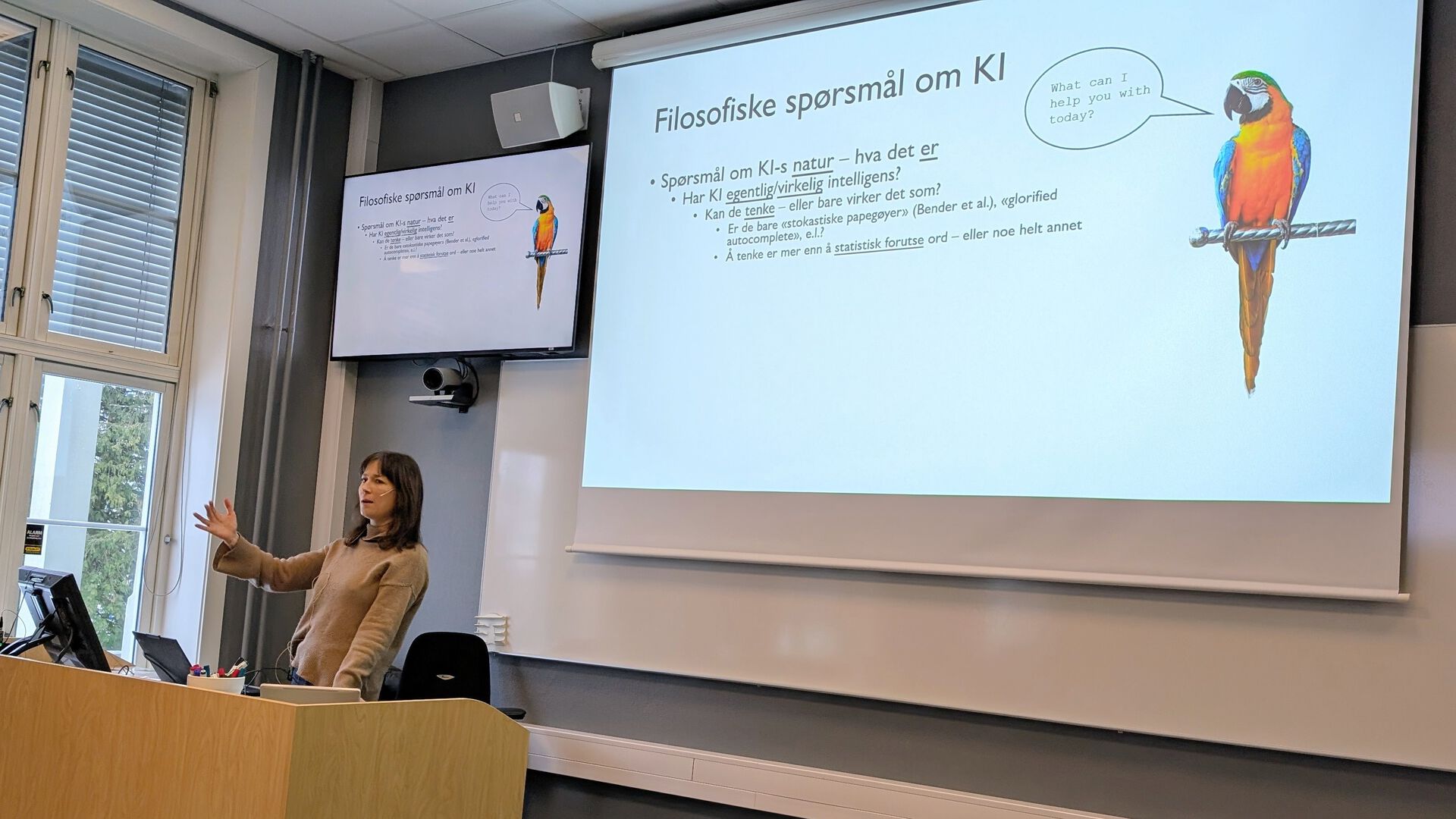 Professor Hedda Hassel Mørch foreleser, på skjermen bak henne er det en powepoint-plakat med overskriften Filosofiske spørsmål om KI og illustrasjon av en papegøye med en snakkeboble med teksten What can I help you with today?