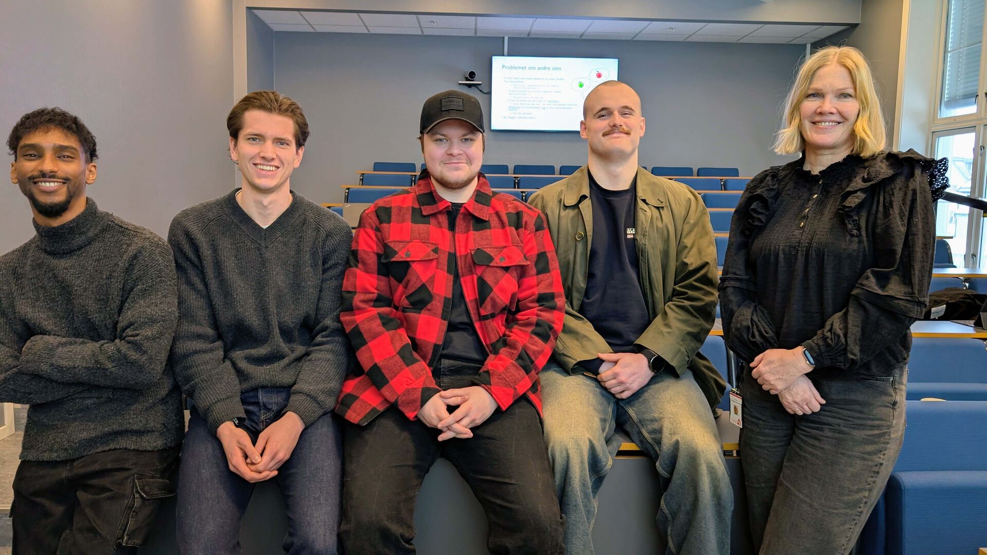 5 studenter oppstilt på rekke i et auditorie. Ser i kamera.