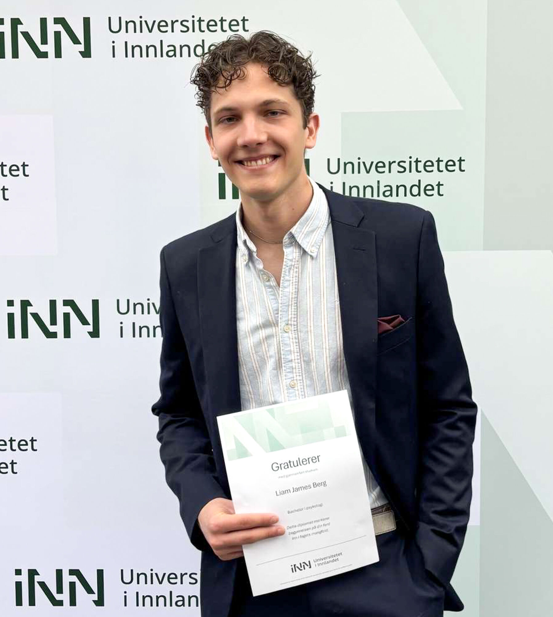 Ung student med vitnemål foran en messevegg med logoen til Universitetet i Innlandet på