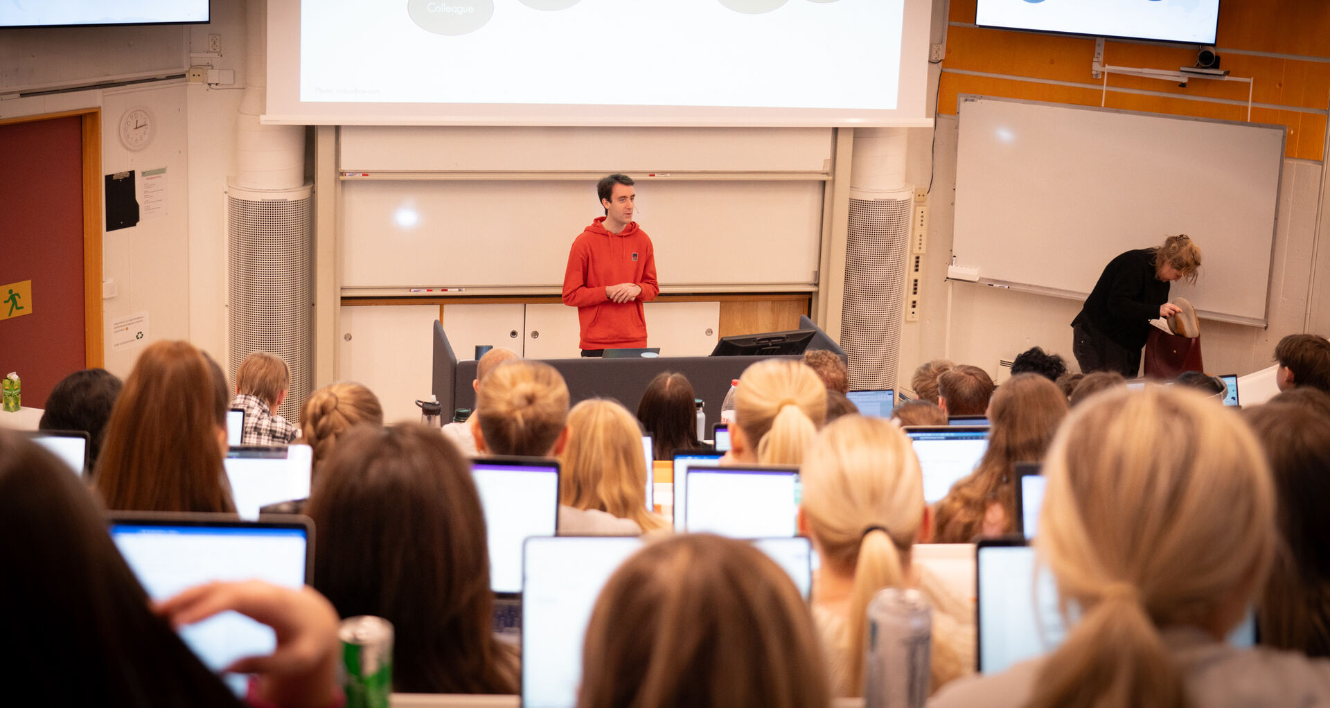Foreleser med rød hettegenser foran masse studenter i et auditorium